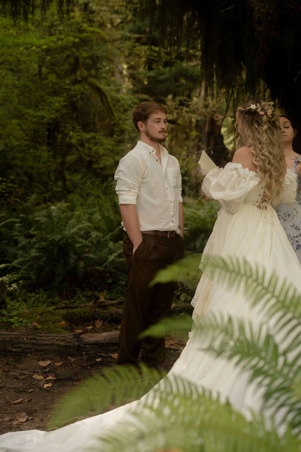 A groom in white shirt and dark pants stands in a lush forest beside a bride in an elegant white gown with floral crown.
