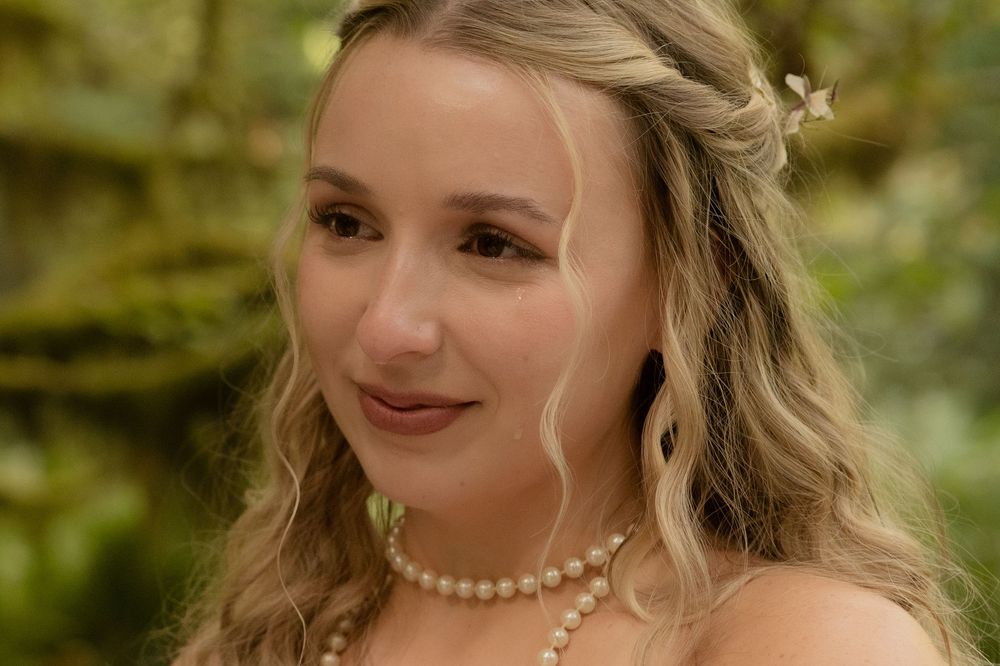 Young woman with blonde wavy hair wearing pearl necklace and hair accessories, posing outdoors in nature.