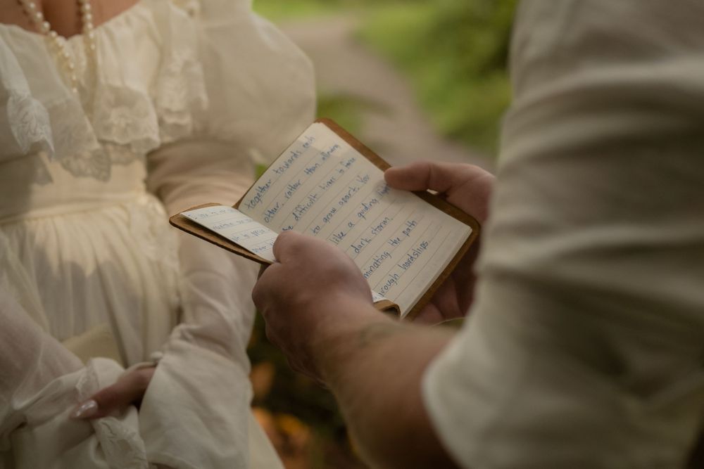 Close-up of hands holding a small handwritten journal, with a person in a vintage white lace dress in the background.