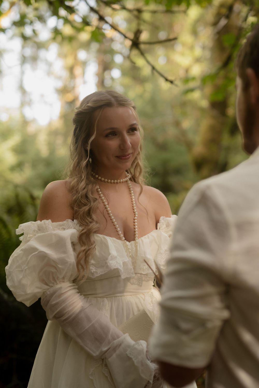 Bride in white off-shoulder gown with pearl necklace smiling in a lush forest wedding ceremony.