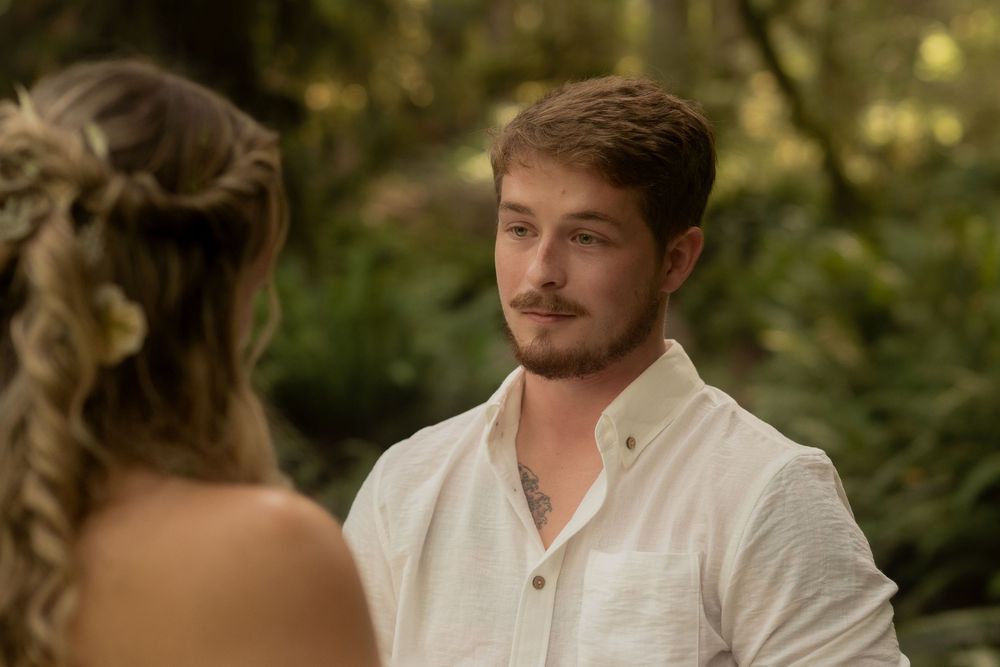 Young man with beard in white shirt stands outdoors in lush green forest setting, facing a woman with braided hair.