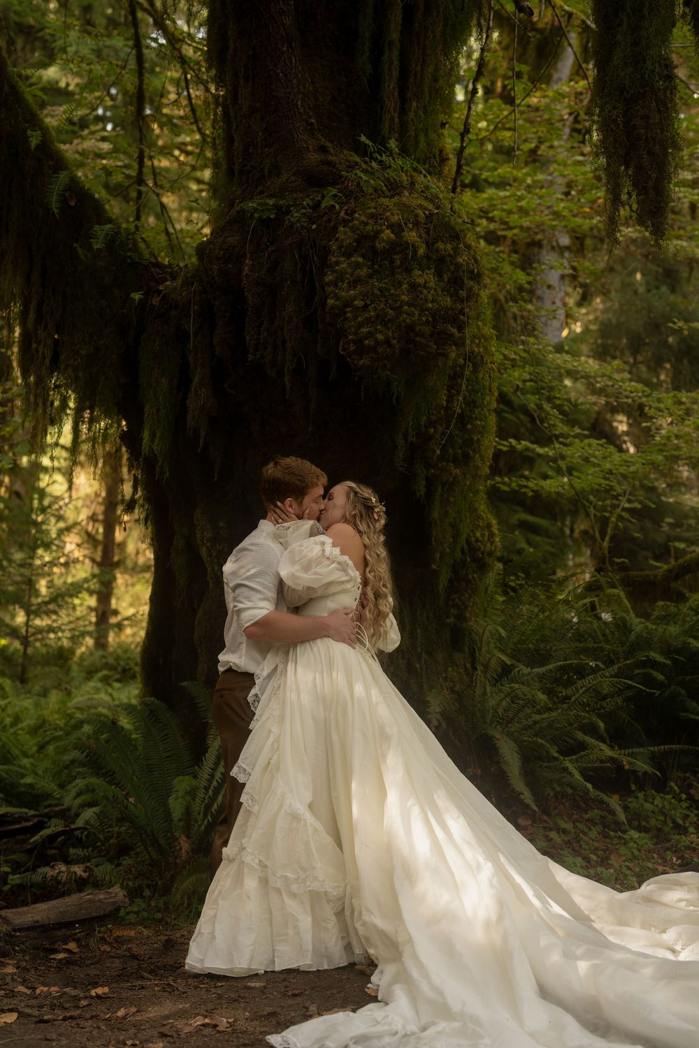 Bride and groom kissing in a lush forest, bride in flowing white gown, surrounded by moss-covered trees and ferns.