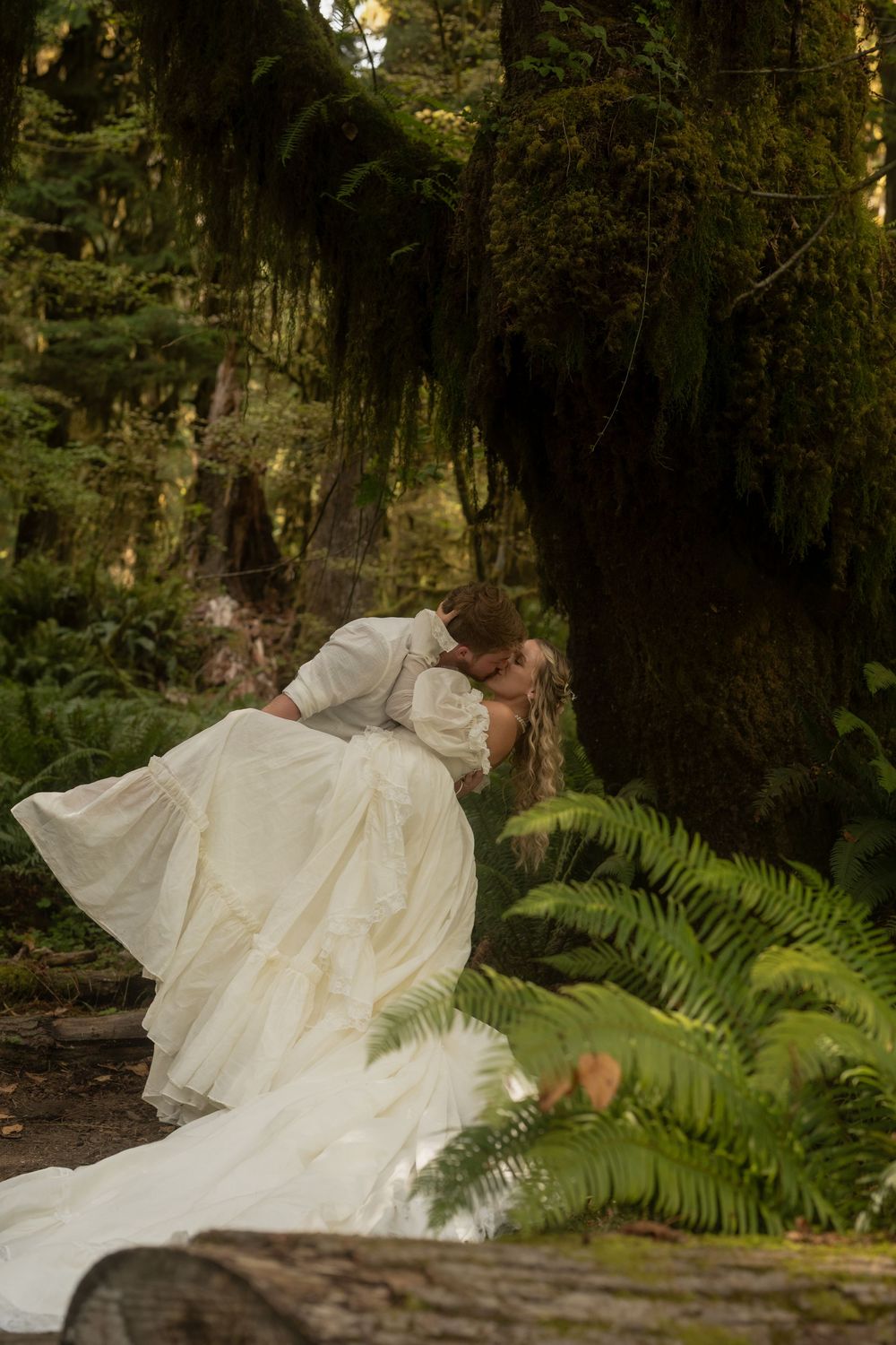 A groom dips his bride in a lush forest setting, her white wedding gown flowing among ferns and moss-covered trees.