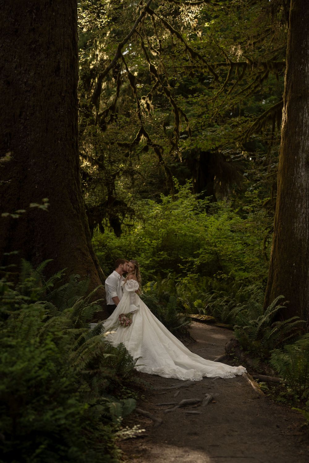 Bride and groom embrace on a forest path surrounded by lush ferns and moss-covered trees.