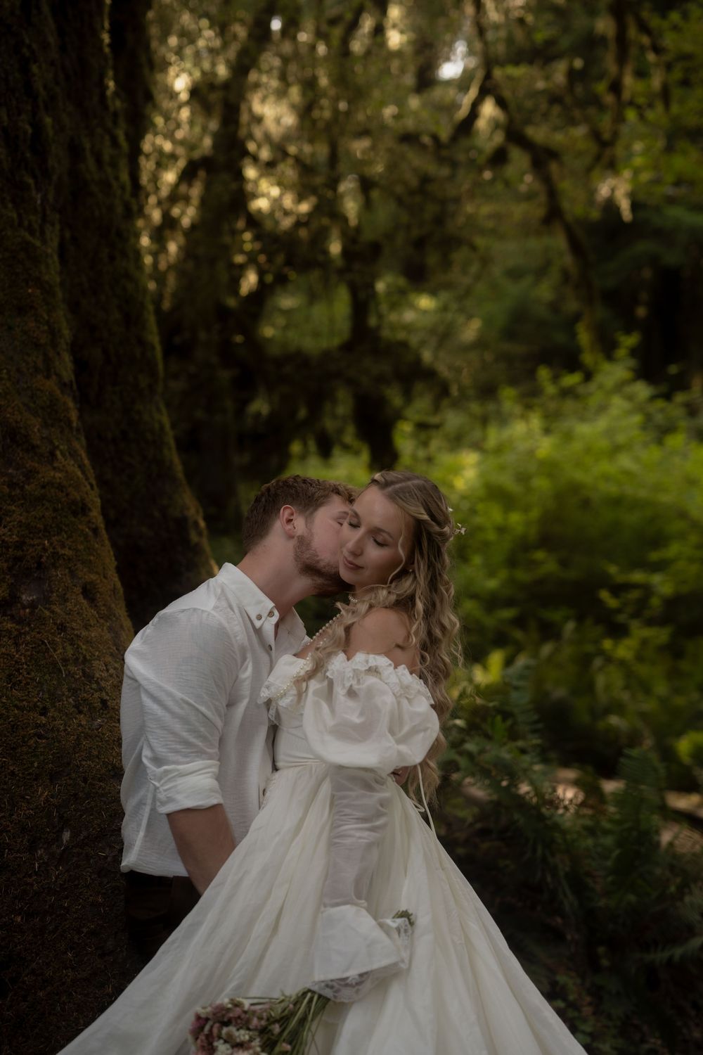 Couple shares romantic moment in forest, groom kissing bride in off-shoulder white gown near mossy trees.