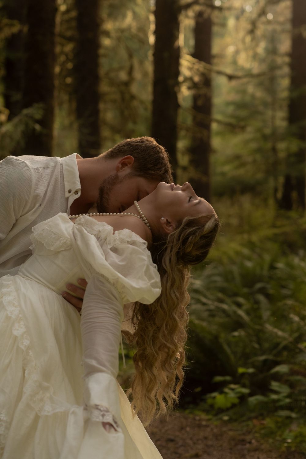 A couple shares a romantic kiss in a lush forest, the bride in a white gown with pearls, groom in a white shirt.