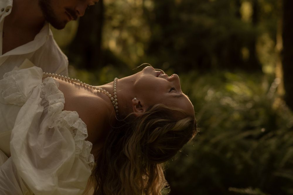 Bride in white ruffled gown with pearl necklace leans back in lush green forest setting.
