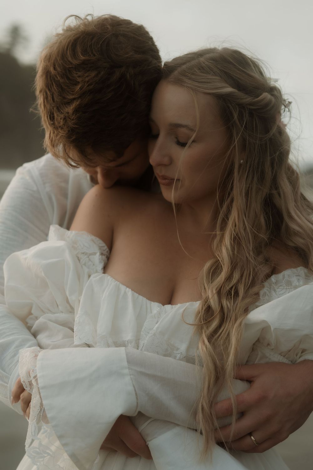 Groom tenderly embraces bride in white off-shoulder gown on a misty beach at dusk.