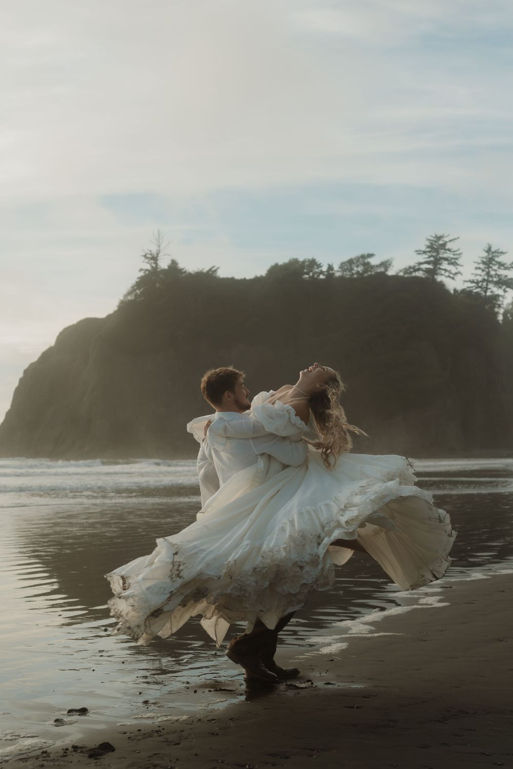 Bride and groom dance joyfully on a misty beach, her white gown flowing in the wind.