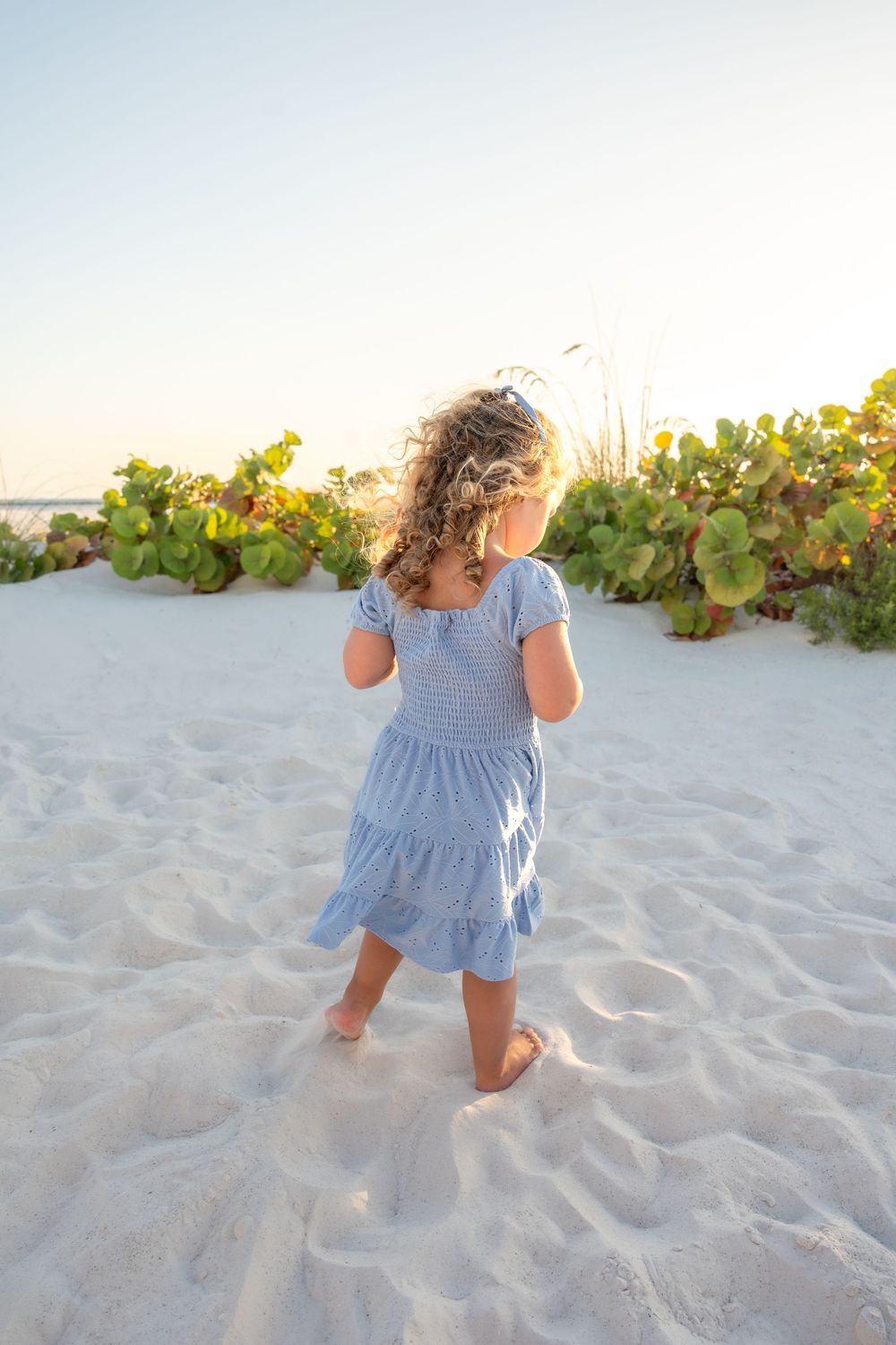 Young girl in blue dress standing barefoot on white sand beach viewed from behind at sunset.