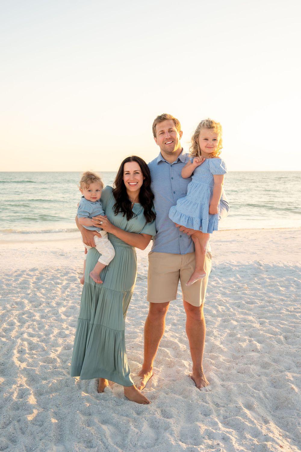 Happy family of four with two young children posing on white sand beach at sunset.