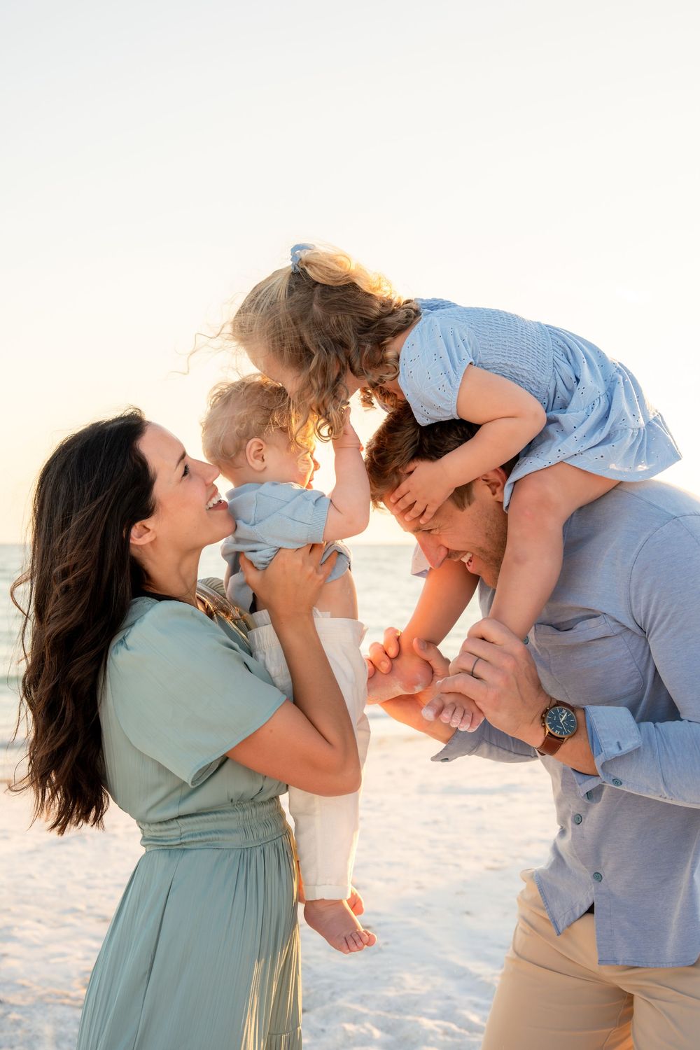 Loving family sharing tender moment at beach sunset, mother holding baby while daughter leans down from fathers shoulders.
