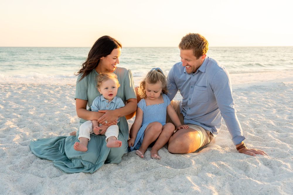 Happy family of four sitting together on white sand beach at golden hour, smiling and enjoying time together.