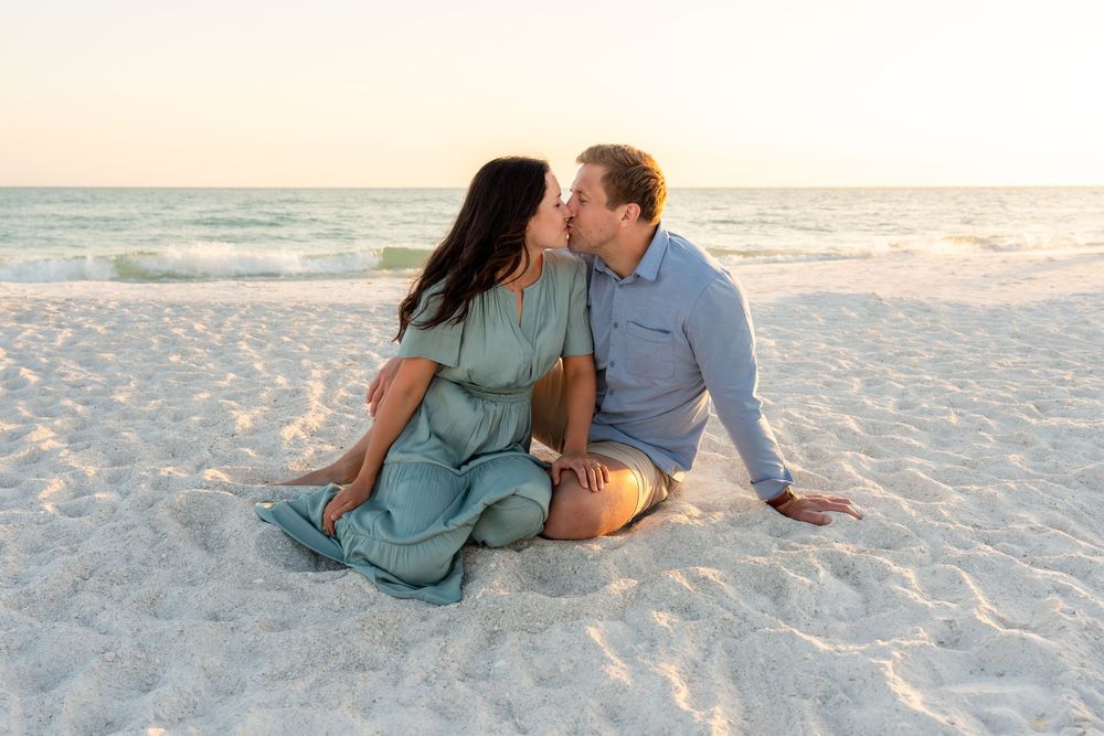 Romantic couple sharing a kiss while sitting on white sand beach during a warm golden sunset.