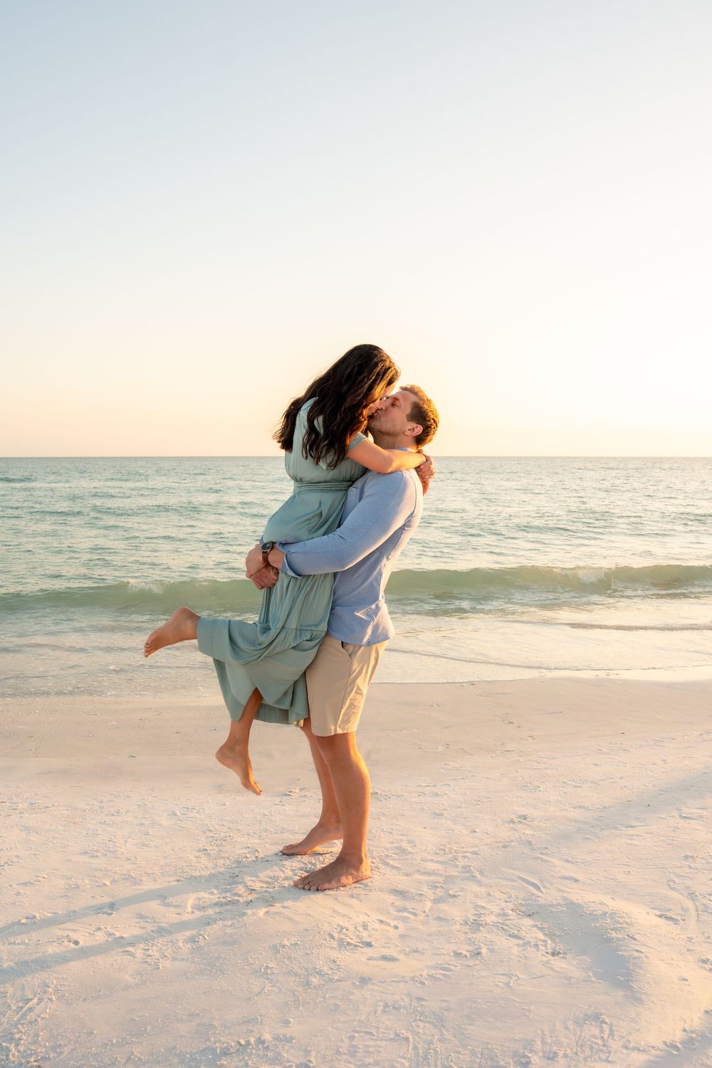 Man lifting and embracing woman on white sand beach at sunset with calm ocean in the background.