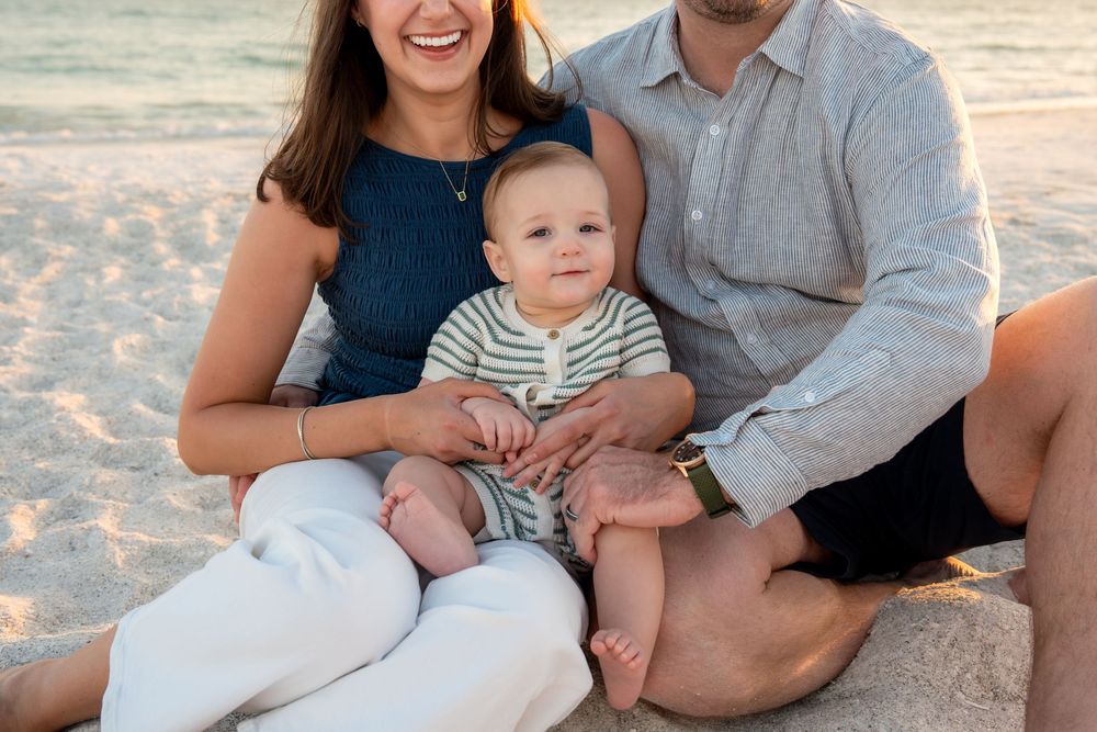 Close-up of family sitting on beach at sunset, parents holding baby in striped outfit.
