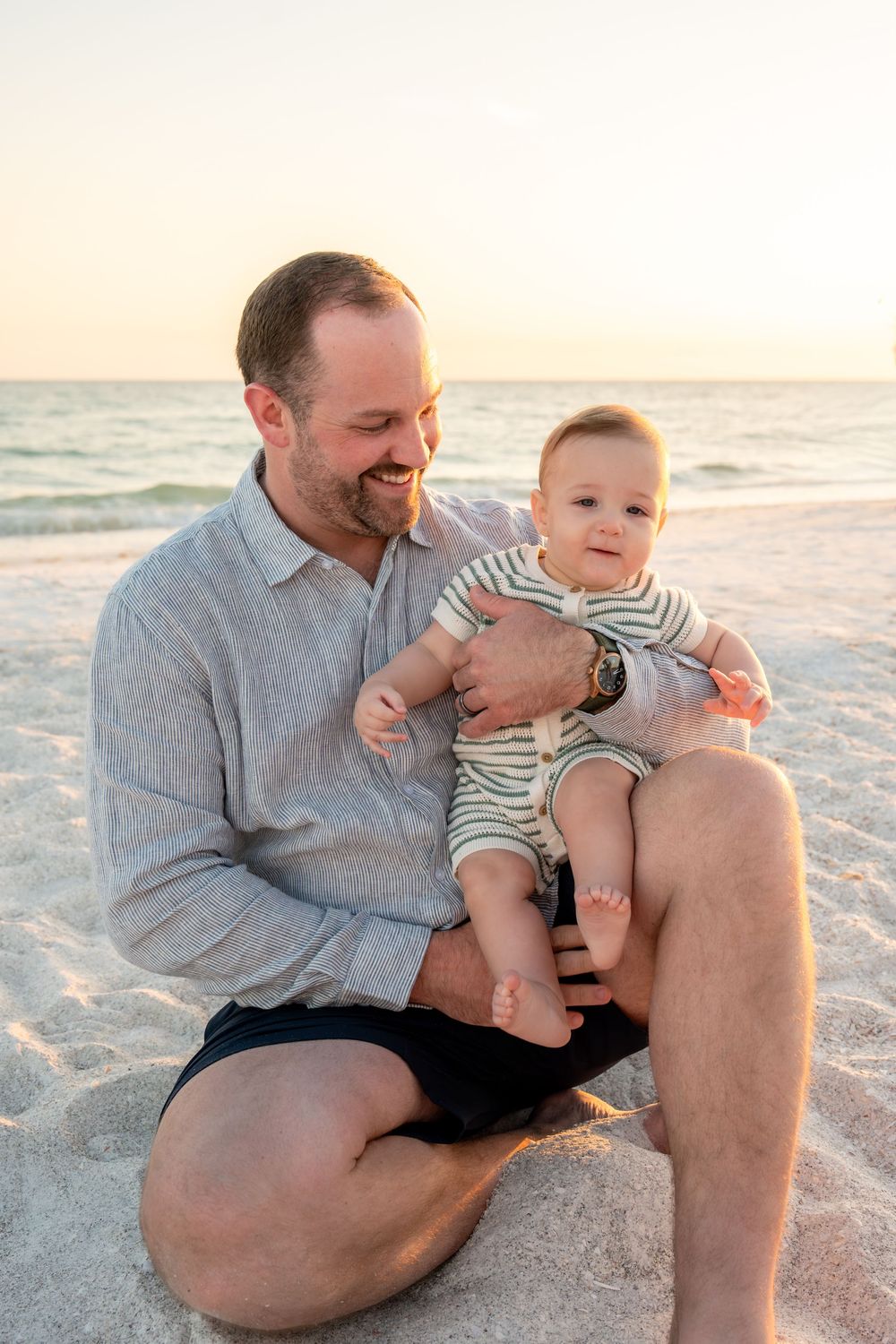 Smiling father sitting on beach sand holding happy baby in striped romper at golden sunset.