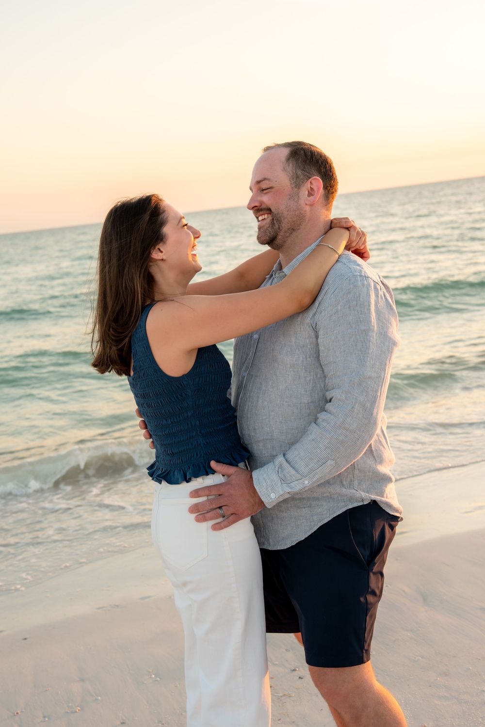 Happy couple embracing and laughing together on beach shoreline during golden sunset.