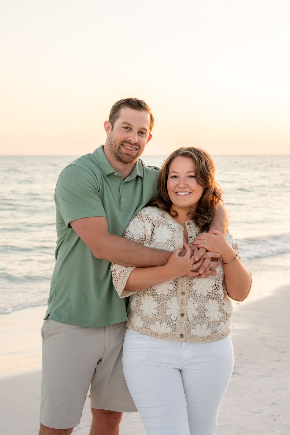Happy couple embracing on sandy beach at sunset, man in green polo hugging woman from behind.