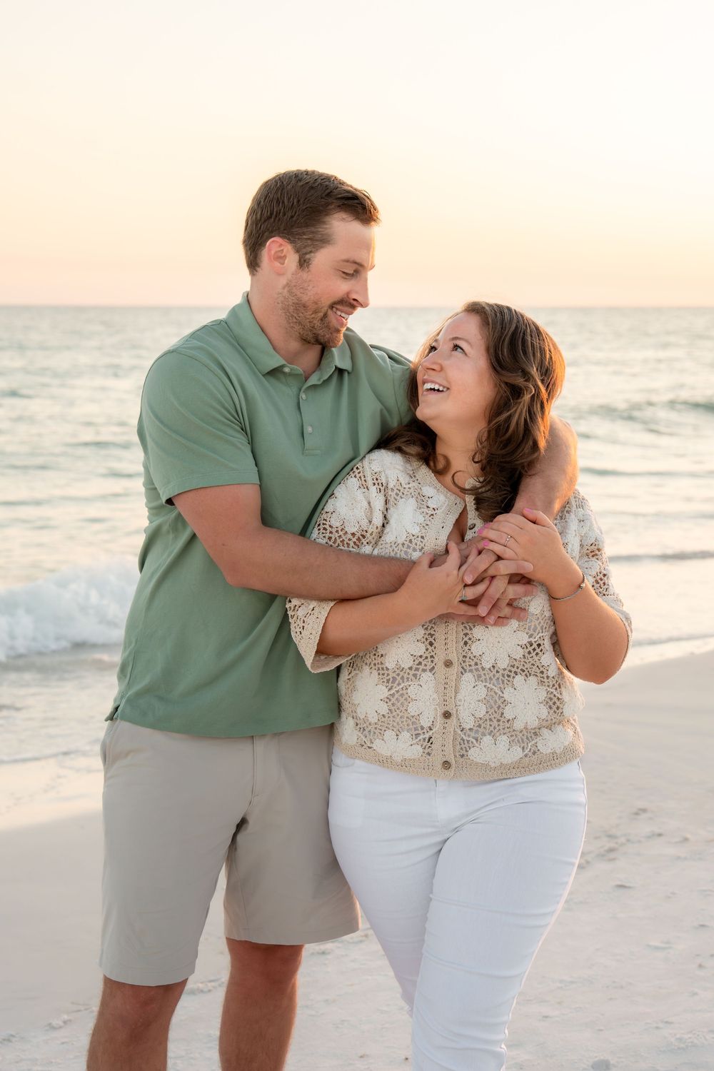 Smiling couple embracing on a beach at sunset, woman in lace top and white pants, man in green polo.