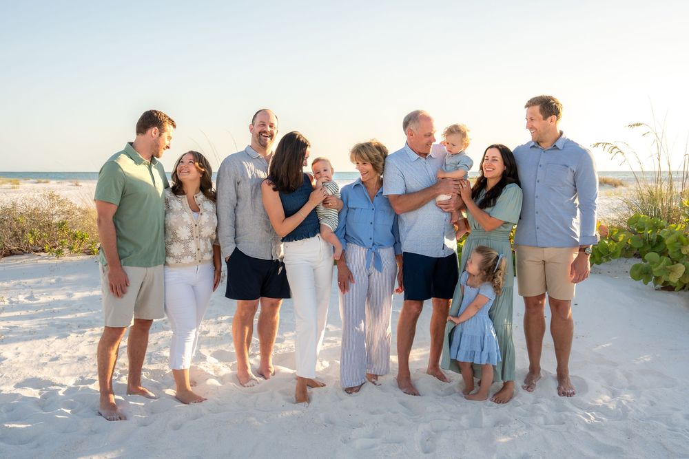 Extended family group smiling together on a bright sandy beach, including young children and grandparents.