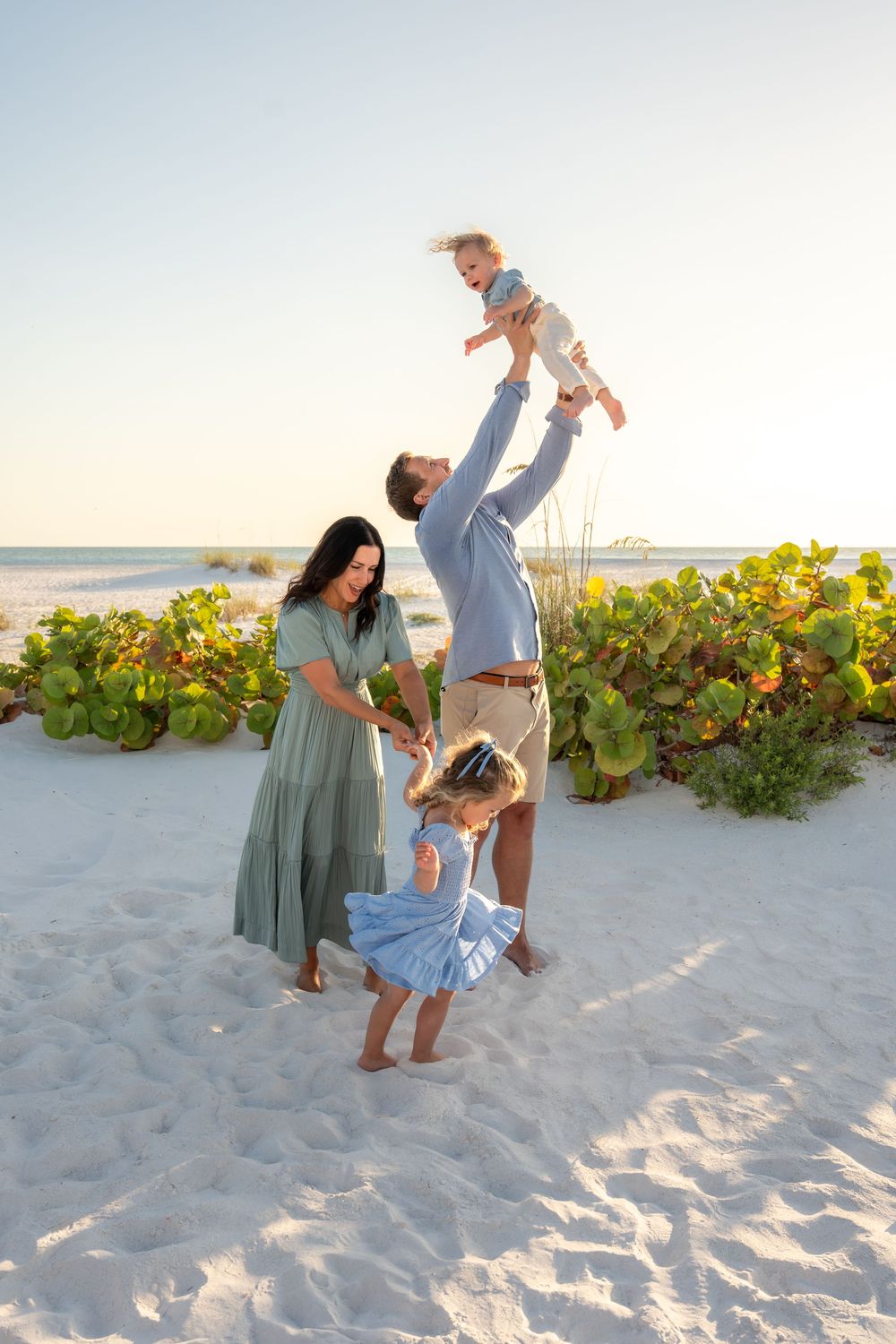 Playful family of four enjoys beach sunset, father lifting baby overhead while mother and daughter laugh on white sand.