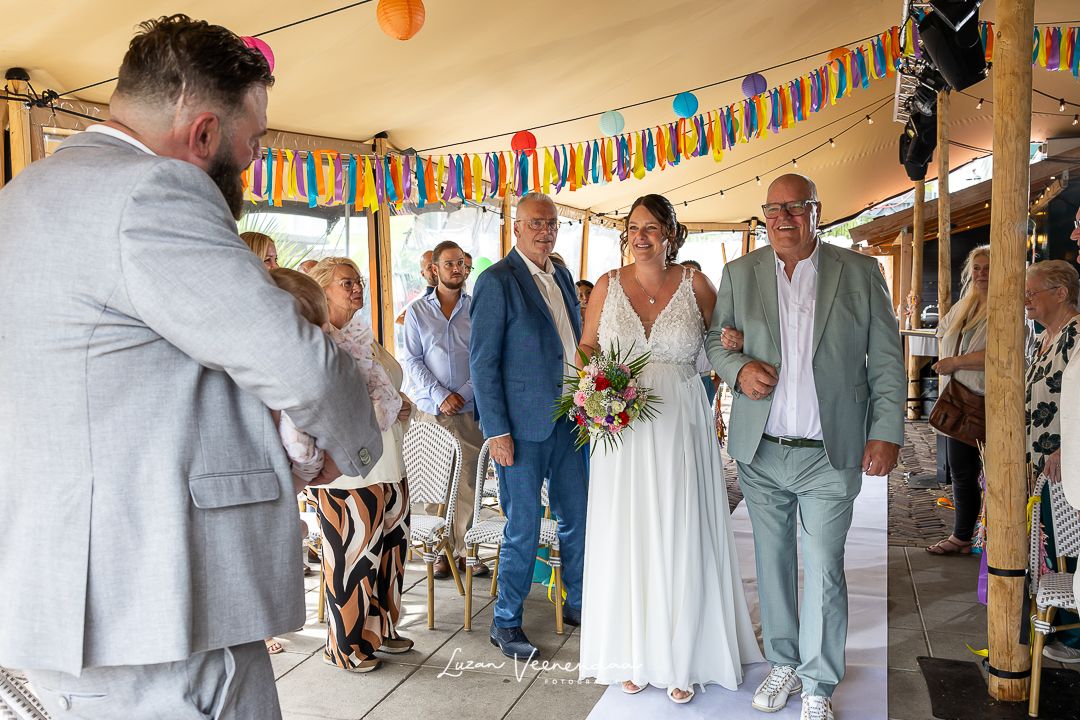 Bruid komt aanlopen met haar vader aan haar arm bij start trouwceremonie  in Zaandam. Bruidegom wacht met baby op zijn arm op zijn bruid.
