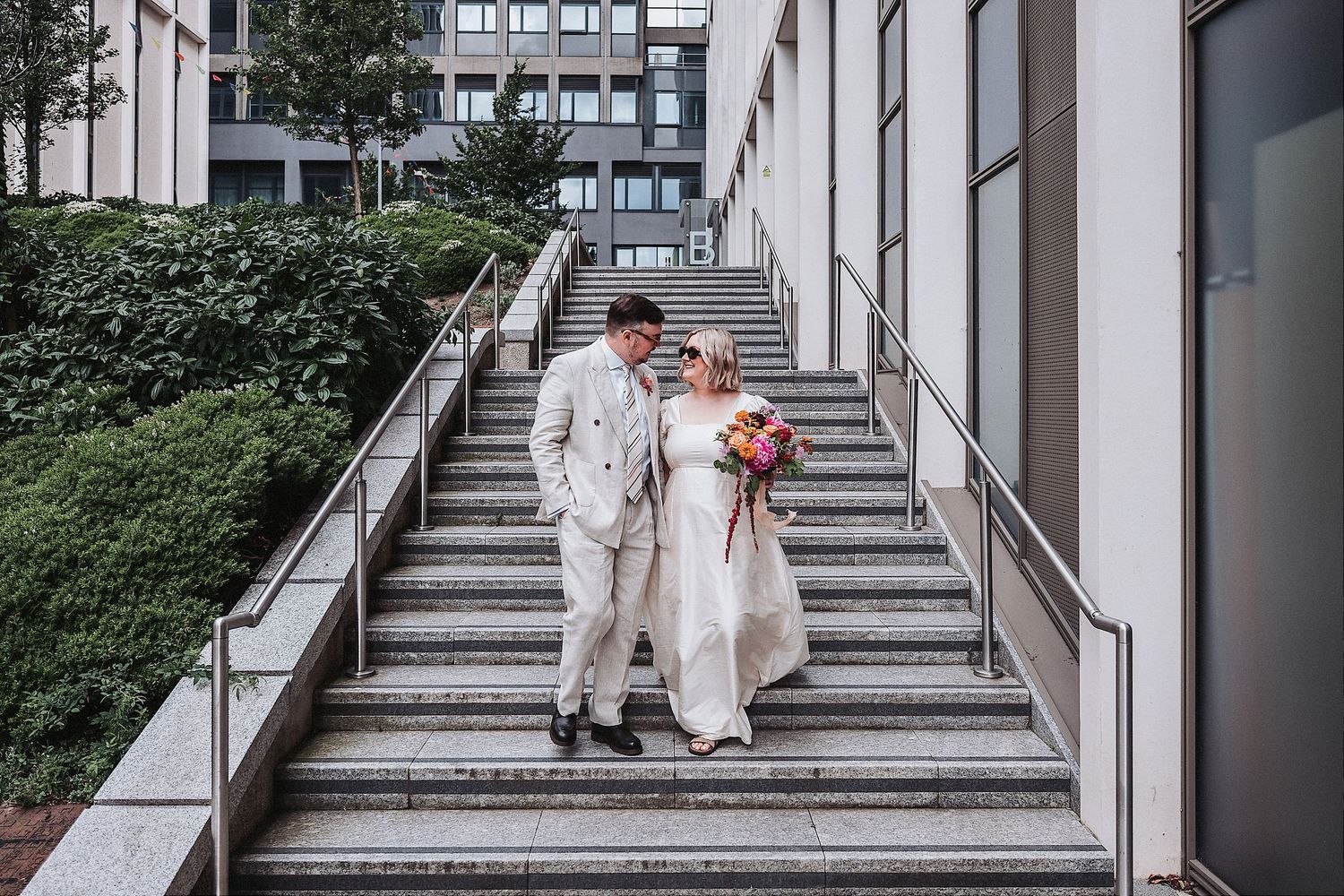 Bride and groom walking down outdoor stairs in urban setting, bride holding colorful bouquet, groom in cream suit.