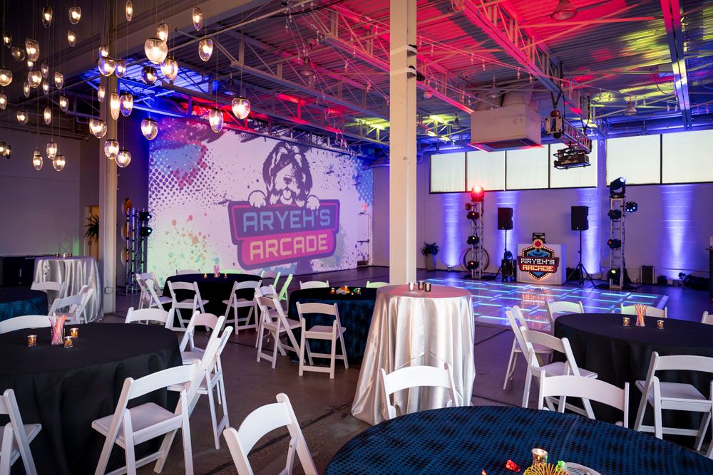 Colorful arcade-themed party venue with white chairs, round tables, string lights, and a vibrant neon gaming backdrop.
