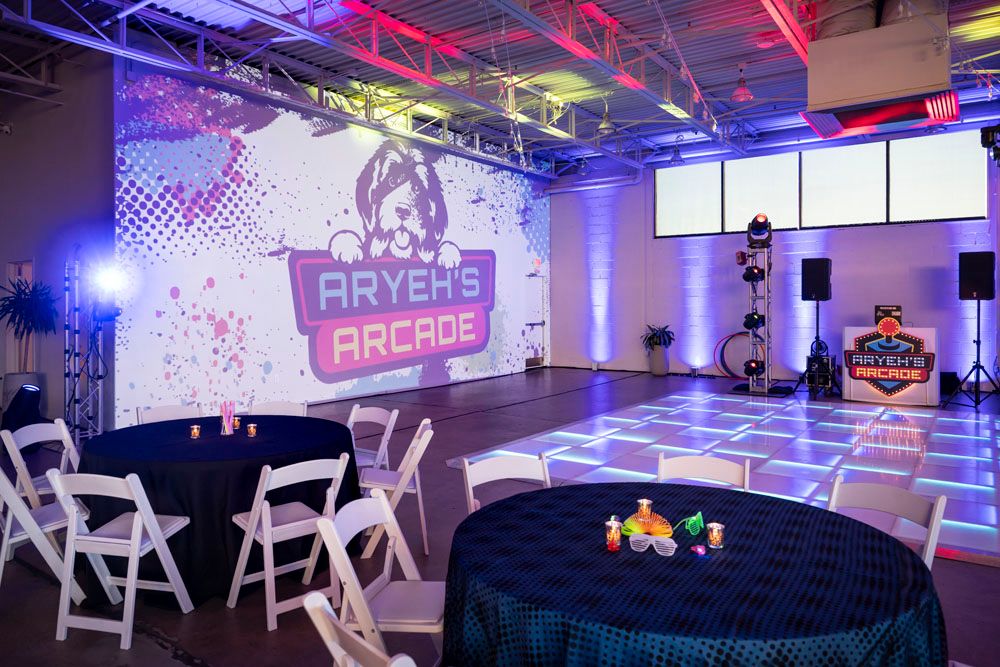 Arcade-themed bar mitzvah venue with LED dance floor, Aryeh's Arcade sign, round tables, and white chairs in colorful lighting.