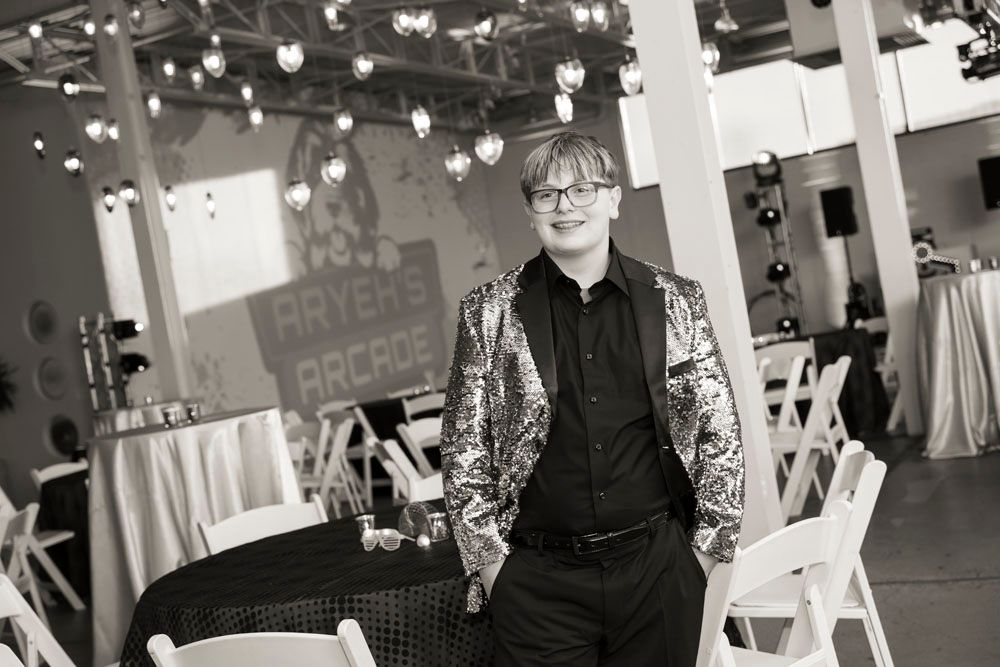 Man in sequined blazer poses confidently at elegant event venue with string lights and decorative signage.