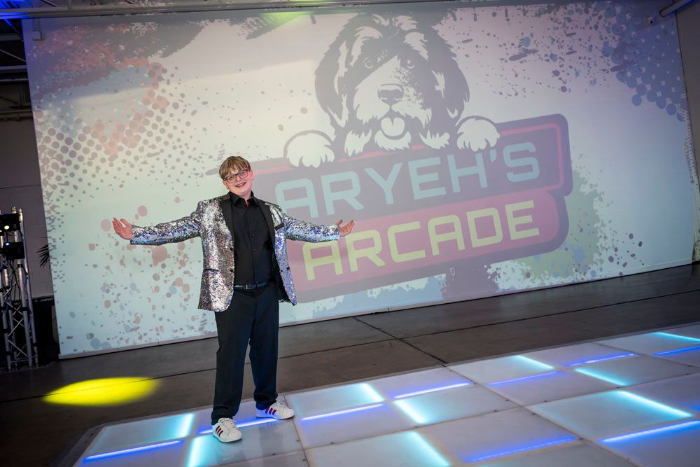 Person posing on a dance floor in front of an Aryeh's Arcade themed backdrop at a celebration event.