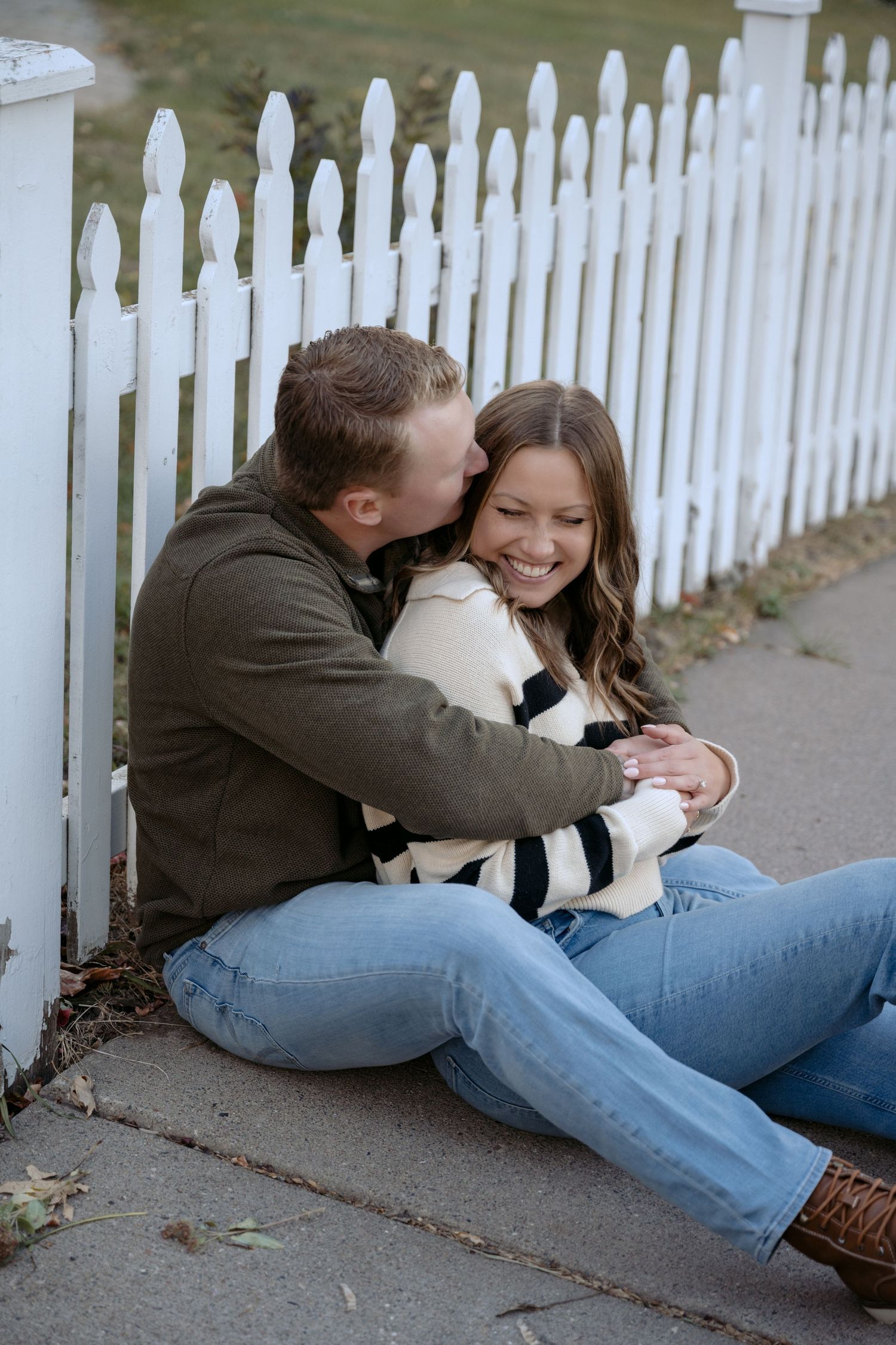 Engaged couple snuggles along a white picket fence for an outdoor suburban engagement session in Stillwater, MN.