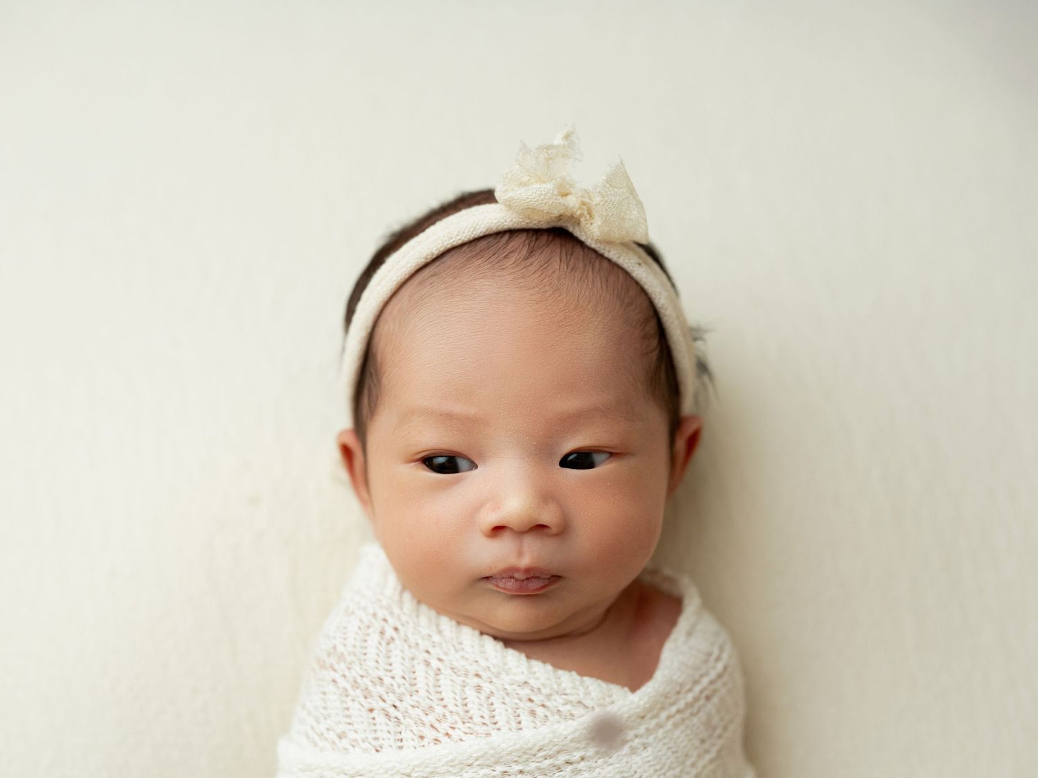Newborn baby girl swaddled in white knit wrap wearing a delicate bow headband, lying on a cream background.