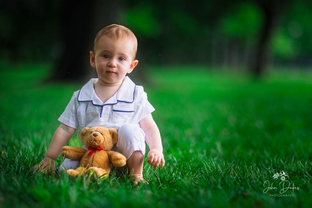 A toddler plays with a teddy bear in lush green grass on a sunny day.