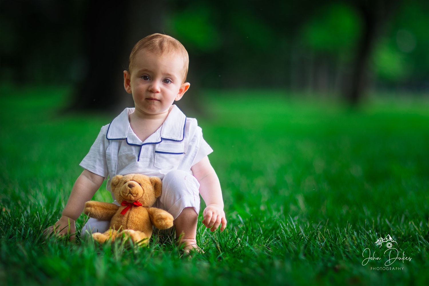 A toddler plays with a teddy bear in lush green grass on a sunny day.