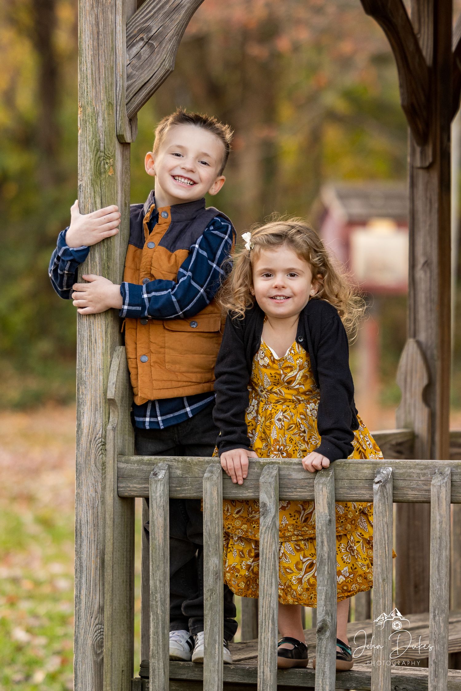Two children smile while playing on a wooden playground structure during autumn.