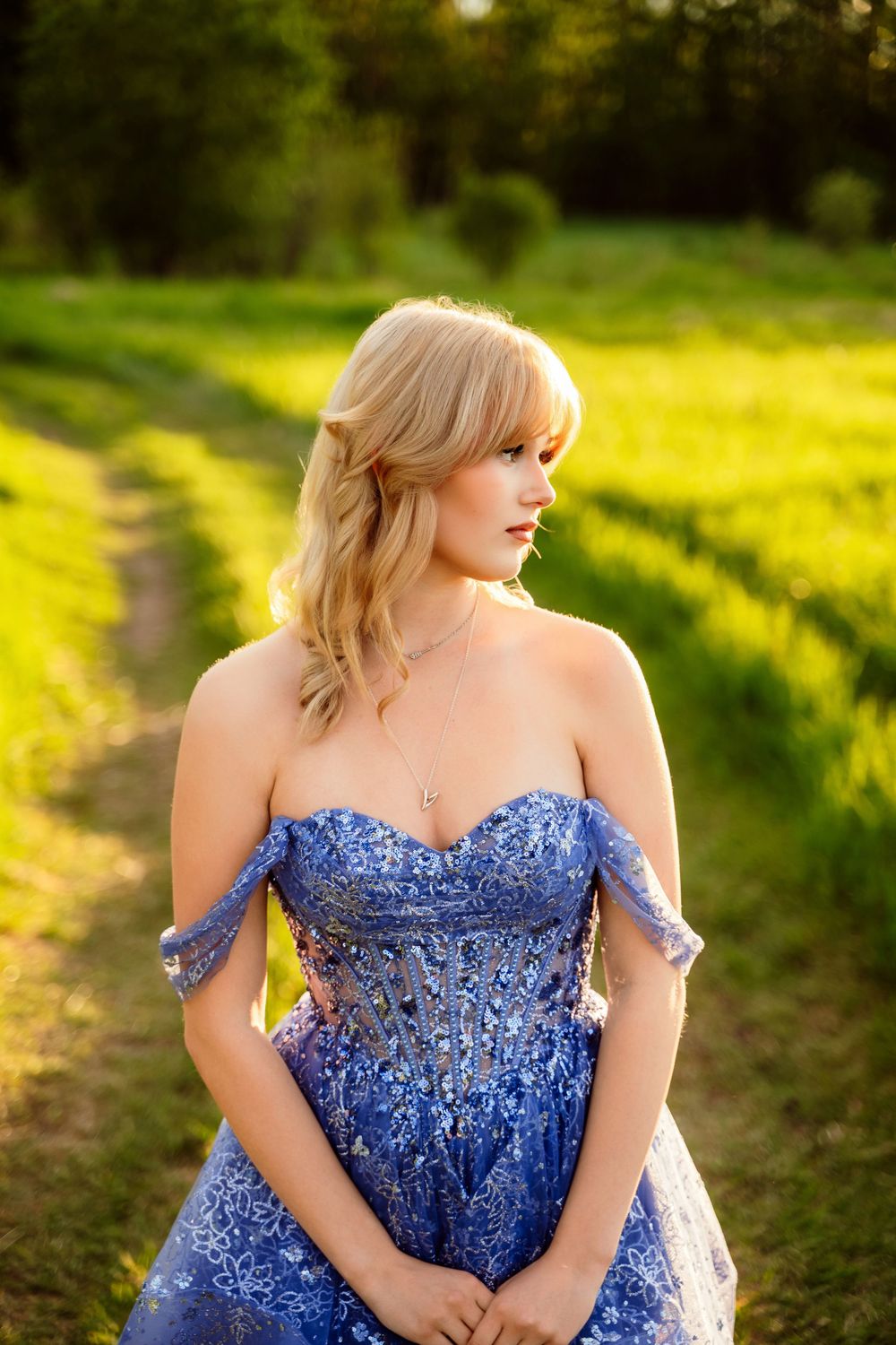 Blonde woman in elegant blue off-shoulder embroidered gown posing outdoors on a sunlit green field path.