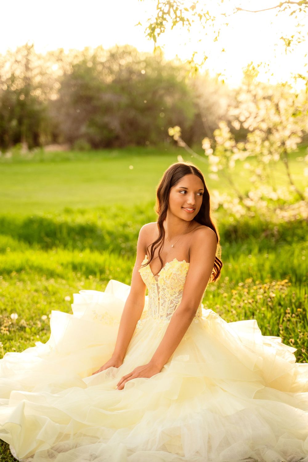 Young woman in elegant ivory ballgown sitting gracefully on green grass with golden sunlight and blooming trees behind her.