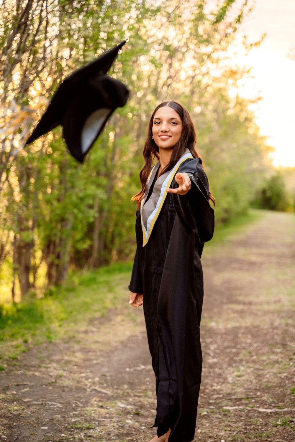 A smiling female graduate in black gown tosses her cap in the air on a tree-lined path at sunset.