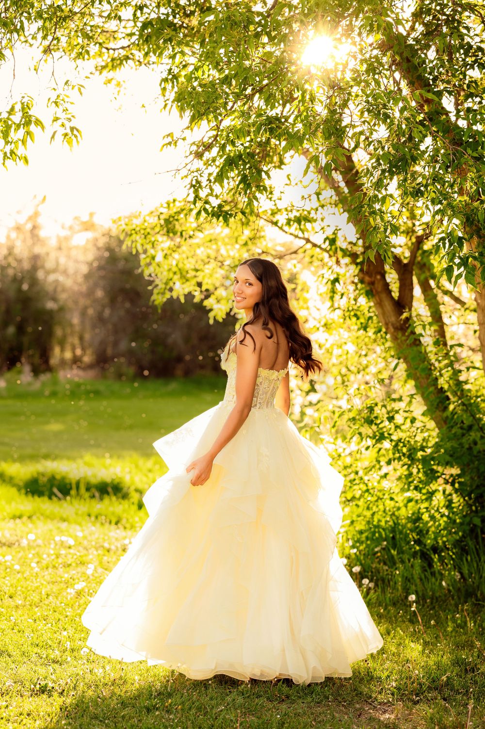 Bride in a flowing white ballgown twirling in a sunlit garden with lush green trees.