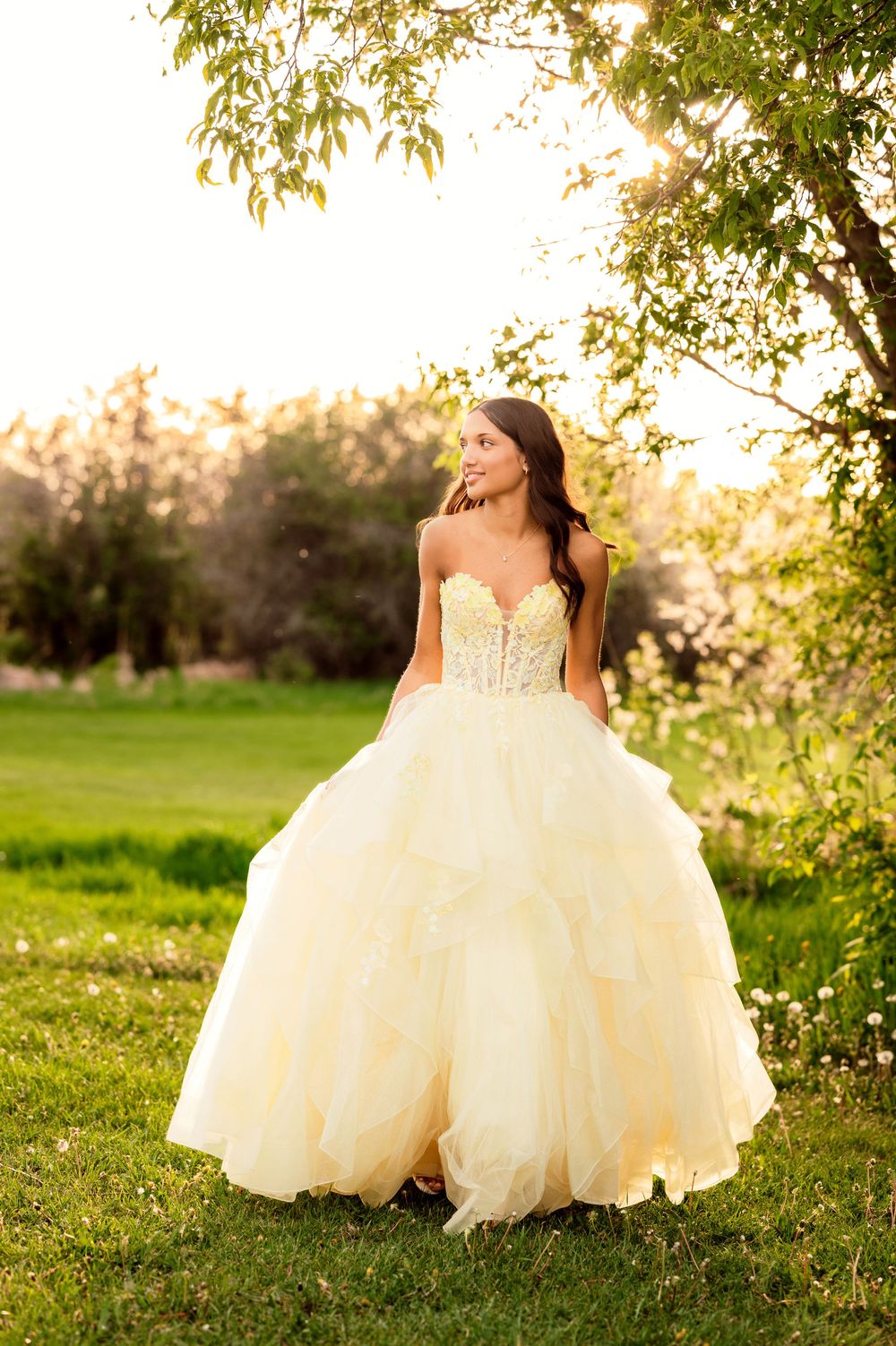 Young woman in a strapless yellow ball gown with ruffled skirt stands outdoors in golden sunlight near green trees.