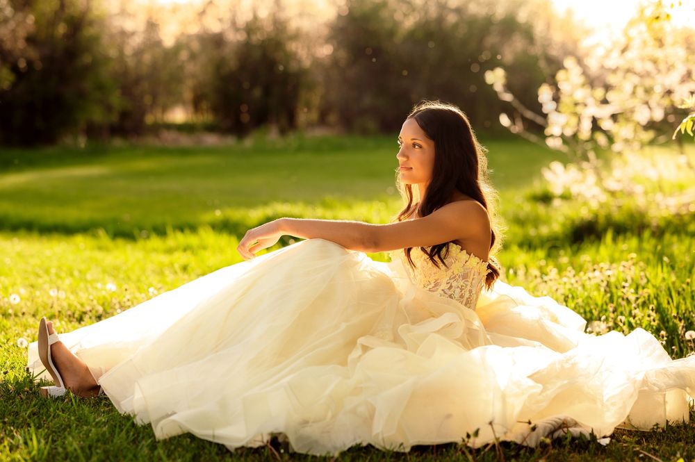 Young woman in elegant ivory ball gown sitting gracefully on green grass in golden sunlight.