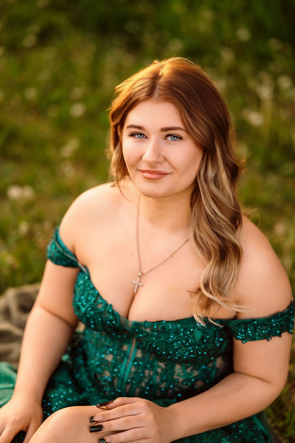 Young woman in green off-shoulder lace dress with cross necklace sitting outdoors in sunlit grassy field.