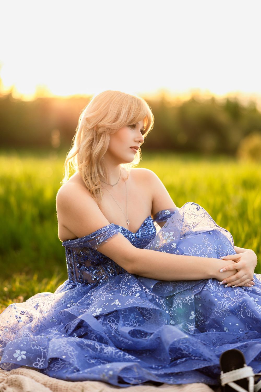 Blonde woman in blue off-shoulder floral gown sitting gracefully in a sunlit green field at golden hour.