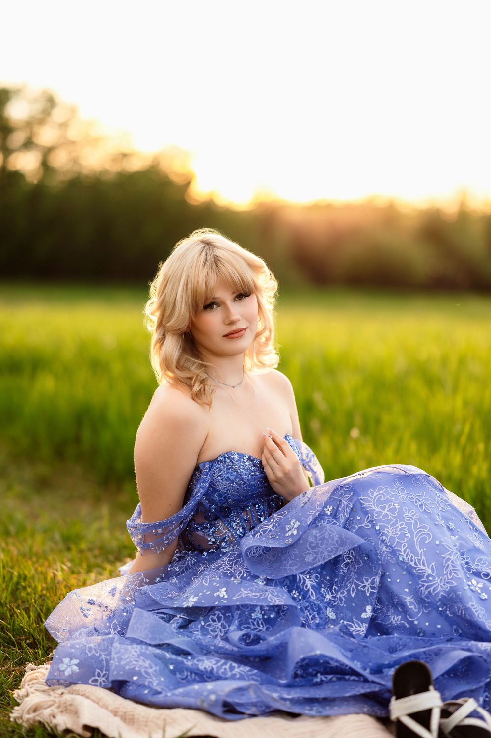 Young blonde woman in a blue ball gown sitting in a sunlit green field at golden hour.