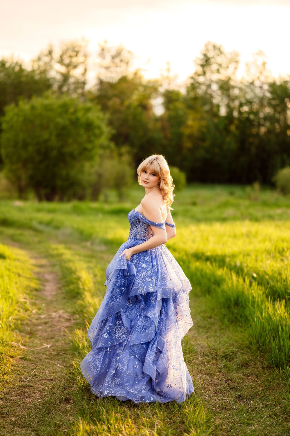 A young woman in a stunning blue off-shoulder ball gown stands on a grassy path surrounded by lush green trees at sunset.