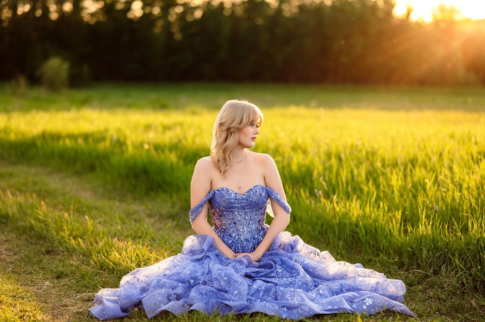 Woman in blue ball gown sitting in sunlit green field at golden hour.