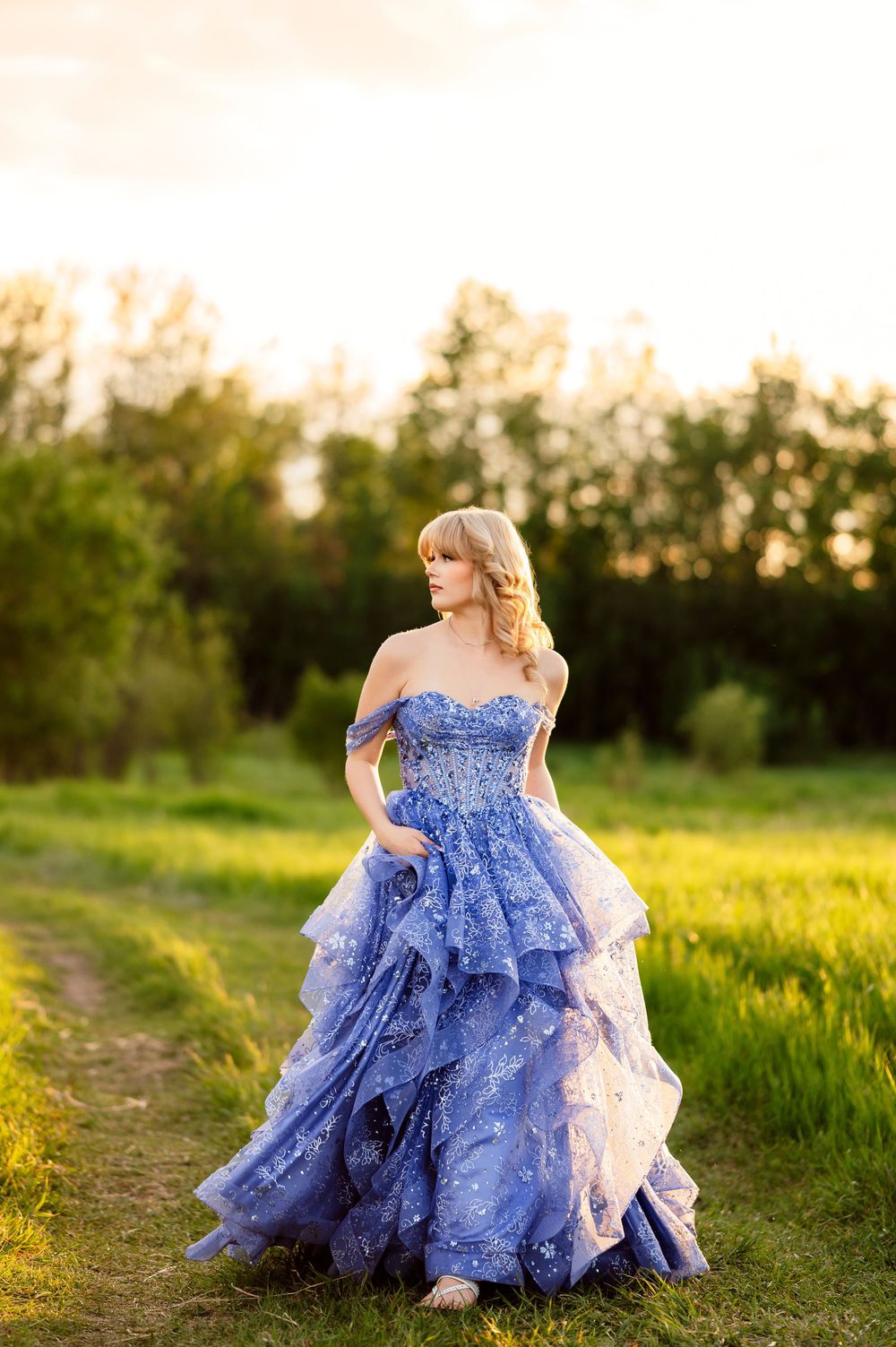 A woman in a stunning blue off-shoulder ball gown stands in a sunlit green field at golden hour.