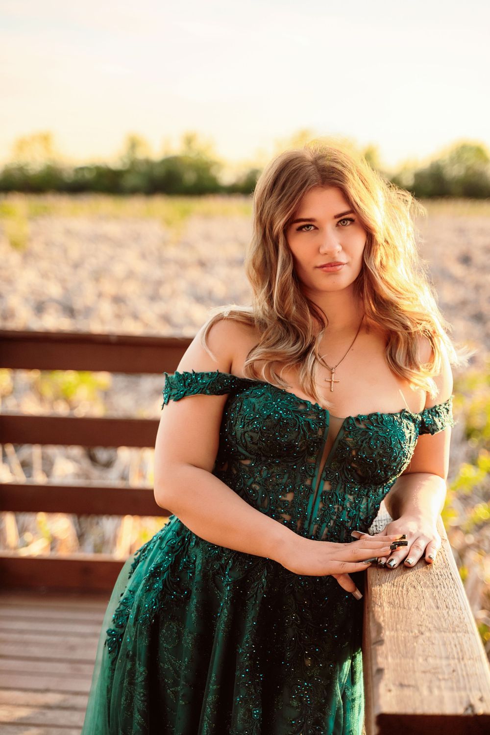 Young woman in elegant emerald green off-shoulder lace gown posing on wooden bridge at golden hour sunset.